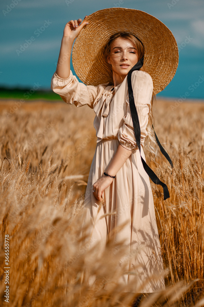 Obraz premium Attractive and beautiful girl in a beige midi dress with a big straw hat, in a wheat field