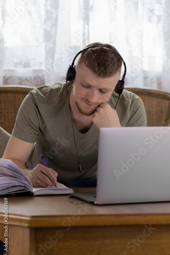 A young student is studying at the university remotely from home using a laptop. Learning in the living room. A man writes information in a notebook with a pen.