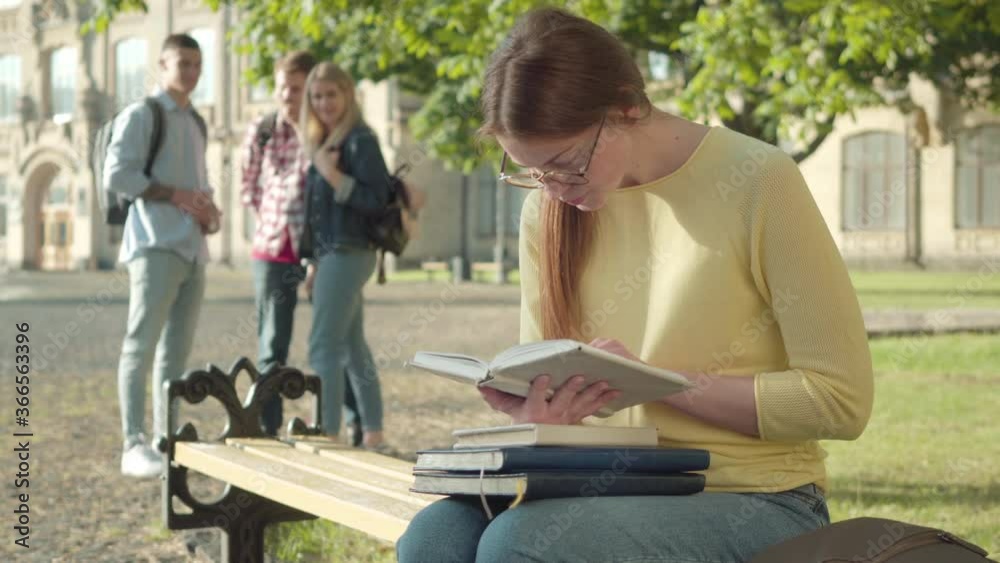Concentrated redhead girl in eyeglasses reading book as blurred groupmates laughing at her at the background. Groupmates bullying shy intelligent young Caucasian student on university campus yard. Concentrated redhead girl in eyeglasses reading book as blurred groupmates laughing at her at the background. Groupmates bullying shy intelligent young Caucasian student on university campus yard.