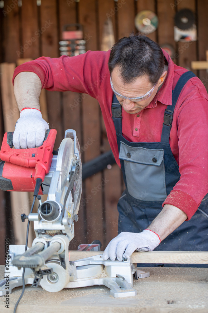 An adult male artisan cuts a wooden plank with a circular saw. Flying sawdust and shavings. Workshop for the manufacture of interior elements for the home. A craftsman works in his carpentry workshop. An adult male artisan cuts a wooden plank with a circular saw. Flying sawdust and shavings. Workshop for the manufacture of interior elements for the home. A craftsman works in his carpentry workshop.
