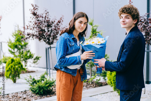 Young woman smiling  her boyfriend meeting her after separation with a bouquet of chamomile. Couple of two students meets in city street outdoor. Surprise  emotions  love and relation concept.