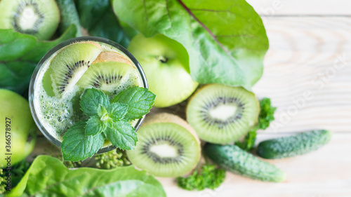 Green smoothie in a glass, decorated with mint and kiwi, with apples, kiwi, cucumbers and parsley in background, selective focus, top view