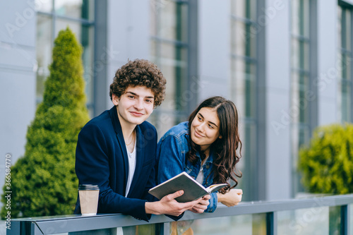 Modest funny boy shy dreamy cute man smiling looking at camera, dreaming or imaging something romantic, holding open book while beautiful girl stands next to him and flirts with him. Urban background