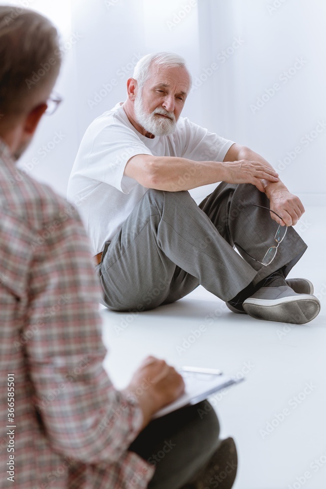 Obraz premium Sad grey senior man sitting on the floor during group psychotherapy for men