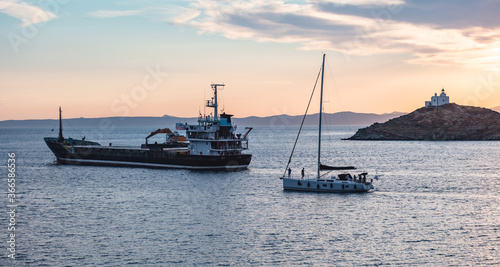 Sailboat and a ship on calm sea at sunset in the Aegean sea, Greece. Lighthouse on a cape at Kea island.