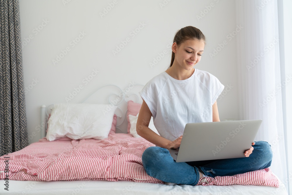 Beautiful girl using laptop and smiling while sitting on bed and studying at home Beautiful girl using laptop and smiling while sitting on bed and studying at home