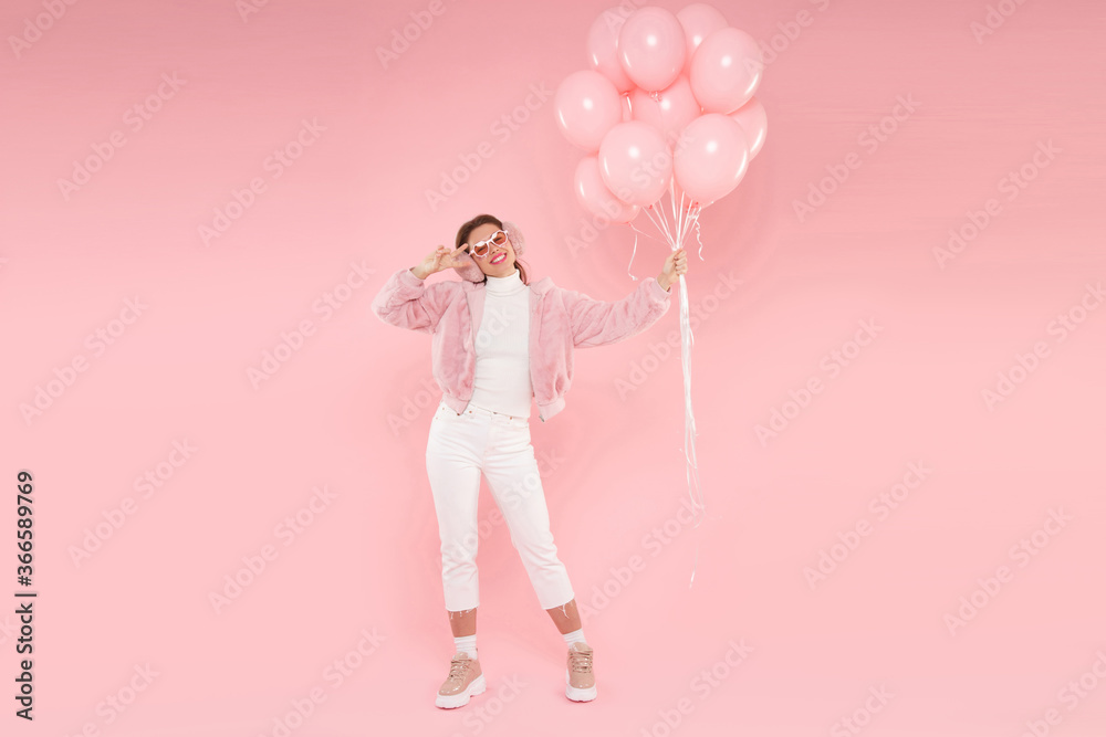 Full length portrait of teen girl wearing fur coat, ear warmers and glasses, holding birthday balloons for party, isolated on pink background