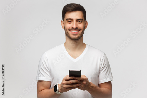 Close-up portrait of young happy smiling male, holding smartphone in both hands, looking at camera with confident face, isolated on gray background © Damir Khabirov