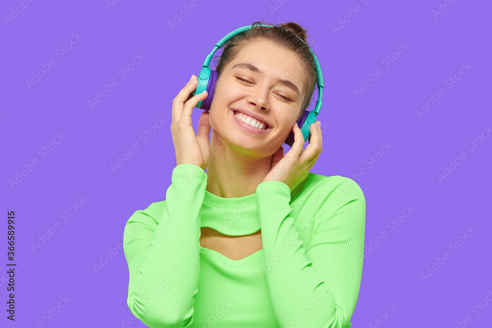 Young happy teen girl wearing neon green long sleeve top, listening to music through wireless headphones with closed eyes, isolated on purple background Young happy teen girl wearing neon green long sleeve top, listening to music through wireless headphones with closed eyes, isolated on purple background