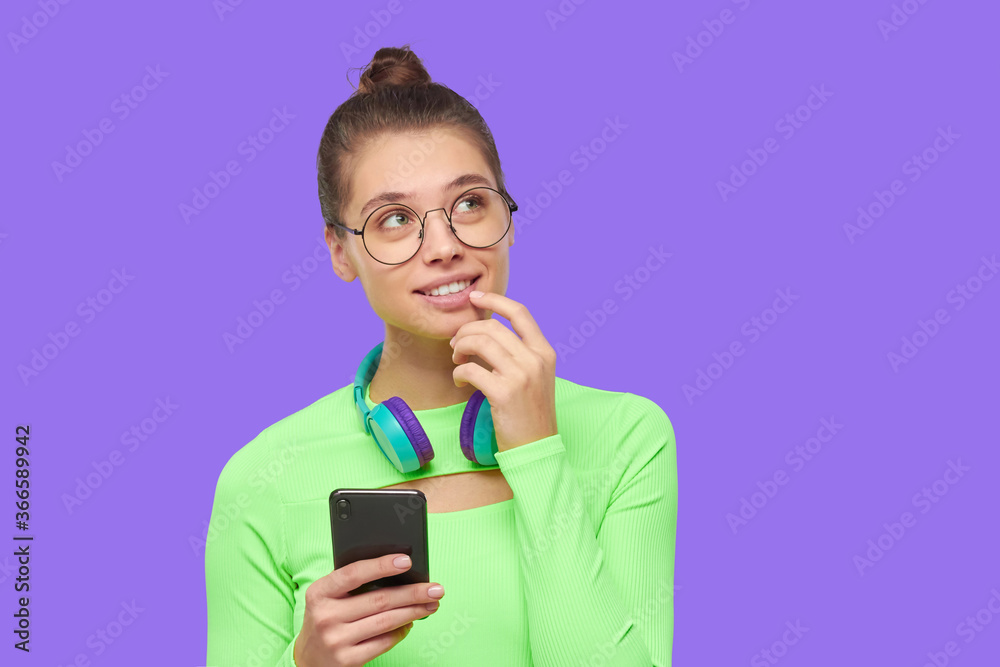Young dreamful girl in glasses, neon green top and with headphones around neck, looking away and dreaming, isolated on purple background Young dreamful girl in glasses, neon green top and with headphones around neck, looking away and dreaming, isolated on purple background