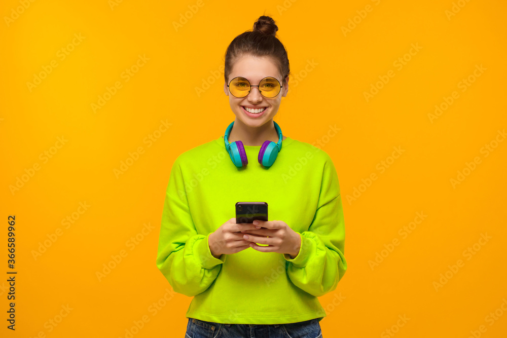 Young teen girl in neon green sweatshirt and glasses, holding smartphone in hands, using app or going to text friends, isolated on yellow background Young teen girl in neon green sweatshirt and glasses, holding smartphone in hands, using app or going to text friends, isolated on yellow background