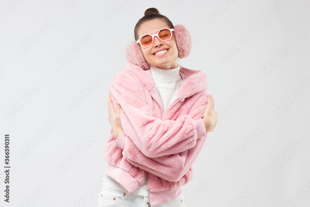 Young girl in pink fur coat, earmuffs and eyeglasses, embracing herself as if dancing and enjoying free time, isolated on gray background Young girl in pink fur coat, earmuffs and eyeglasses, embracing herself as if dancing and enjoying free time, isolated on gray background