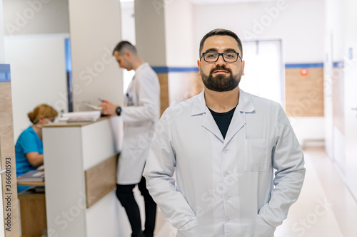 Portrait of bearded doctor in white scrubs. Medical worker stands in hospital corridor.