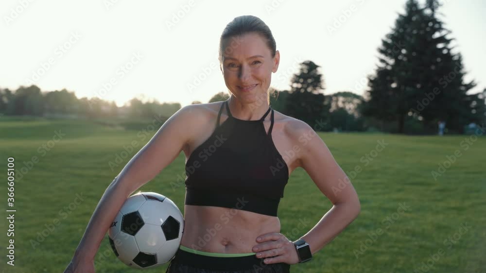 Ready to play. Portrait of attractive sporty woman holding soccer ball in hand, looking at camera and smiling while standing in green field on a summer morning Ready to play. Portrait of attractive sporty woman holding soccer ball in hand, looking at camera and smiling while standing in green field on a summer morning