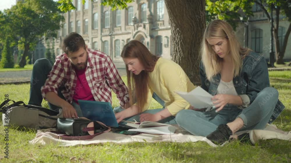 Three active university students studying outdoors on sunny day. Portrait of interested young Caucasian men and women talking and checking documents. Education, lifestyle, intelligence.