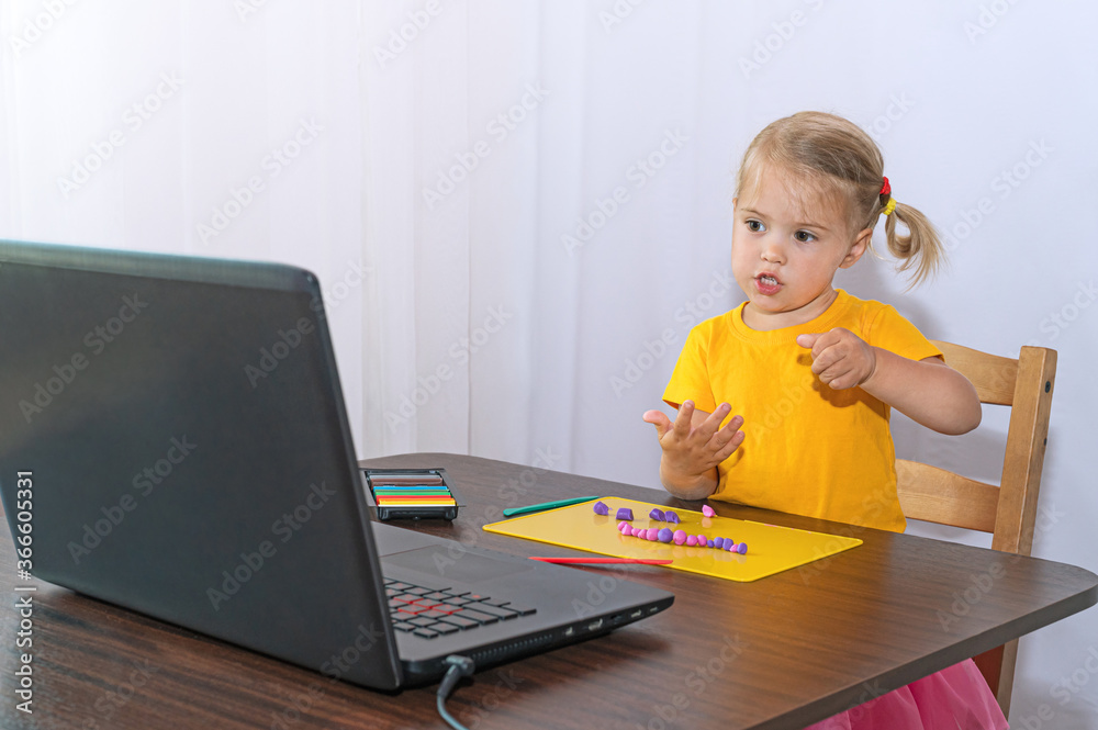An interested little girl looks at her laptop and sculpts out of plasticine. Interested girl in an online lesson next to her laptop. An interested little girl looks at her laptop and sculpts out of plasticine. Interested girl in an online lesson next to her laptop.