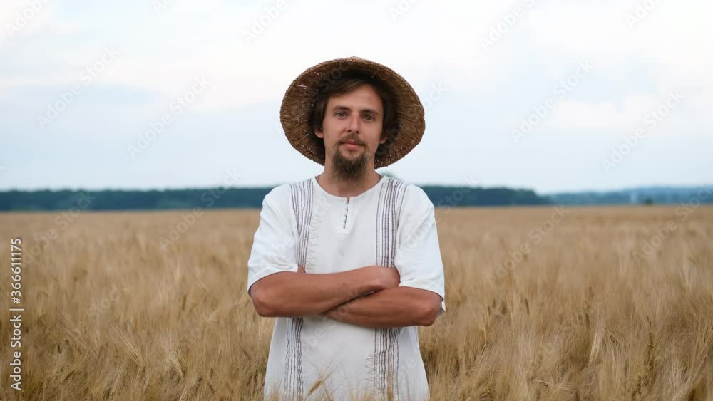Portrait of a brutal young farmer with a beard and straw hat standing on a wheat yellow field and smiling at the camera and folding his arms Portrait of a brutal young farmer with a beard and straw hat standing on a wheat yellow field and smiling at the camera and folding his arms