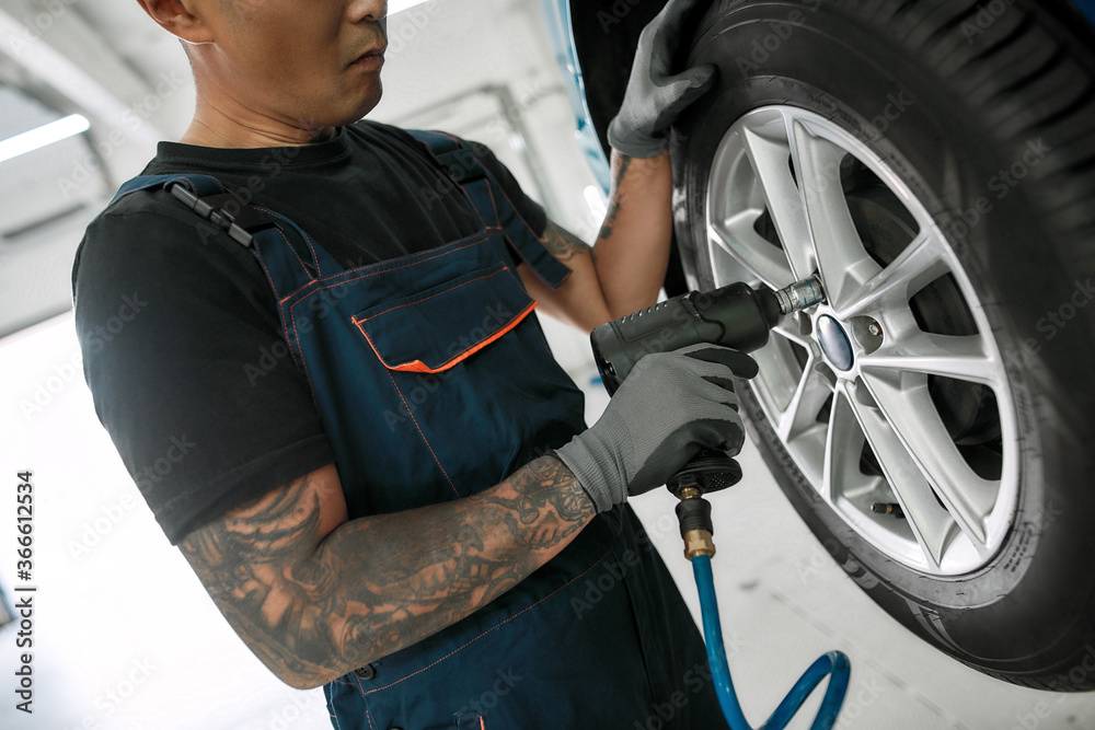 Obraz premium Cropped shot of male mechanic screwing or unscrewing car wheel of lifted automobile by pneumatic wrench at auto repair shop