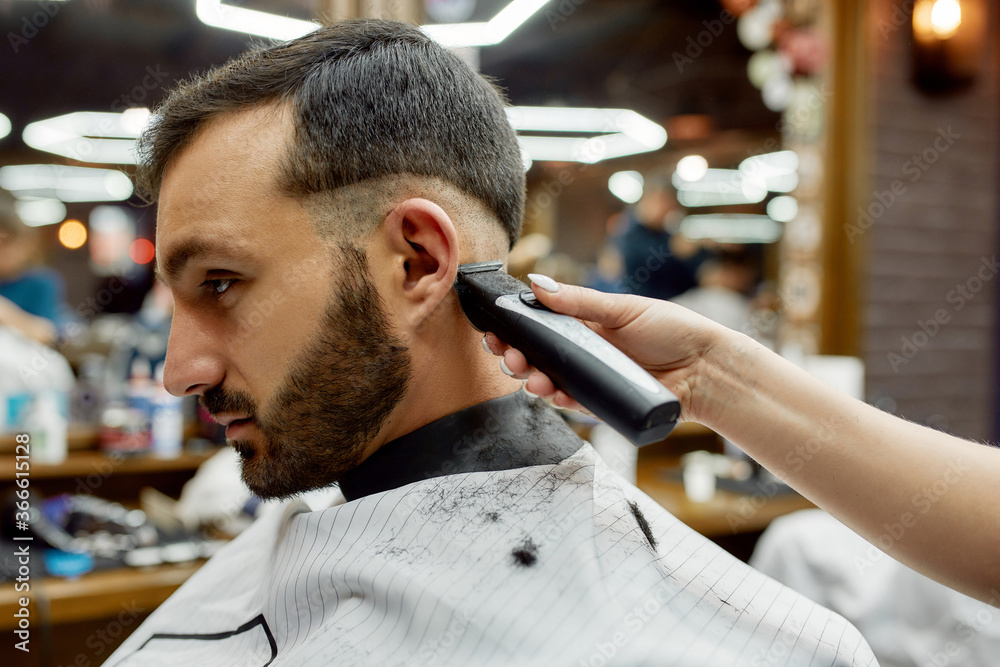 Side view of a young handsome bearded man getting new trendy haircut while visiting modern barbershop. Barber using professional electric hair clipper Side view of a young handsome bearded man getting new trendy haircut while visiting modern barbershop. Barber using professional electric hair clipper