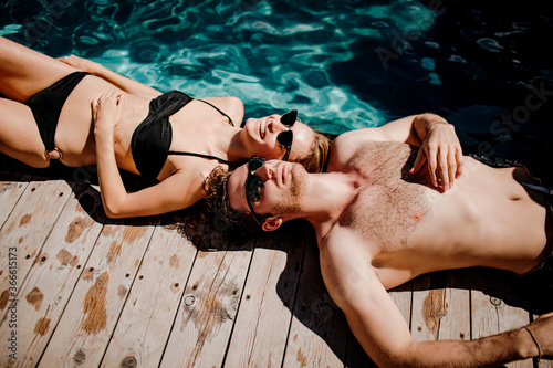 Young hot couple resting together. Side view of cheerful man and woman lying at swimming pool and enjoying relax rest time. Sunbathing after swimming.