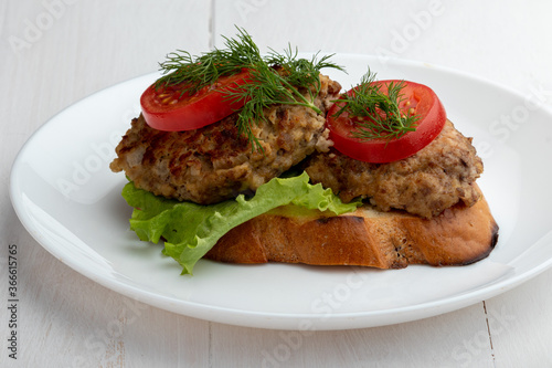 sandwich with cutlet, lettuce and tomato slices on a white plate on a white wooden background