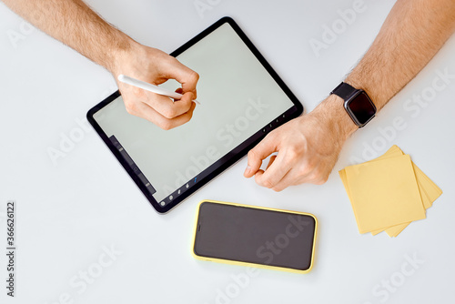 Man works on tablet with stylus next to phone and paper for notes 