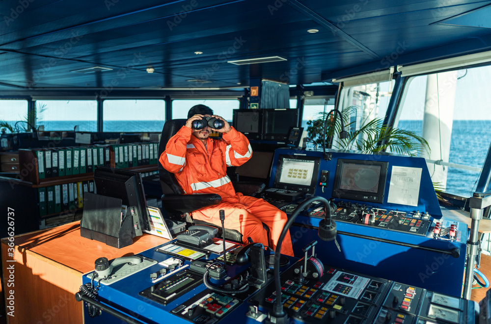 Fototapeta premium Filipino deck Officer on bridge of vessel or ship wearing coverall. He is looking through binoculars. COLREG navigaton watch at sea
