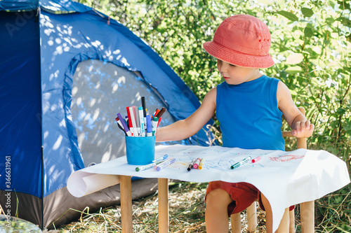 a boy in red Panama sits at a children's table and draws markers on a white watman in nature near the camping tent