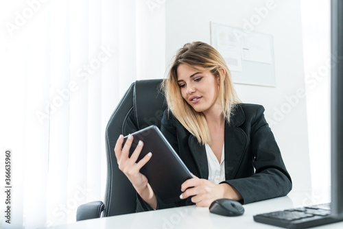 Successful blonde businesswoman sitting at work desk and holding tablet.