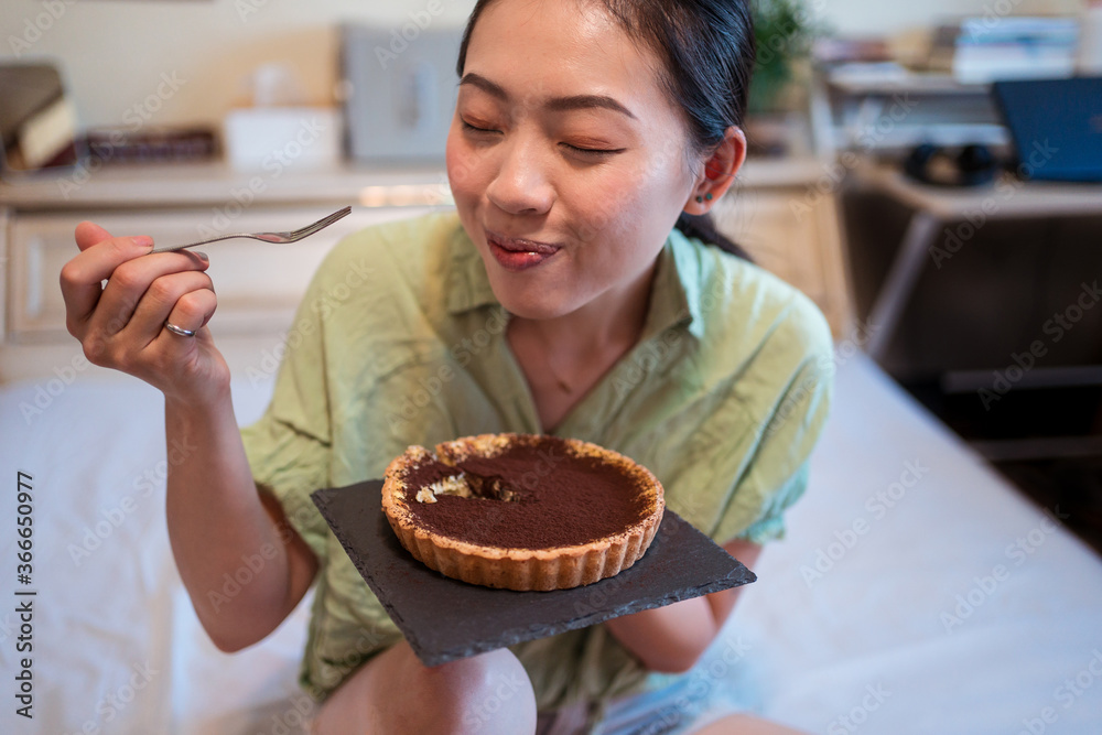 Young pretty Asian woman enjoying delicious chocolate tart on bed