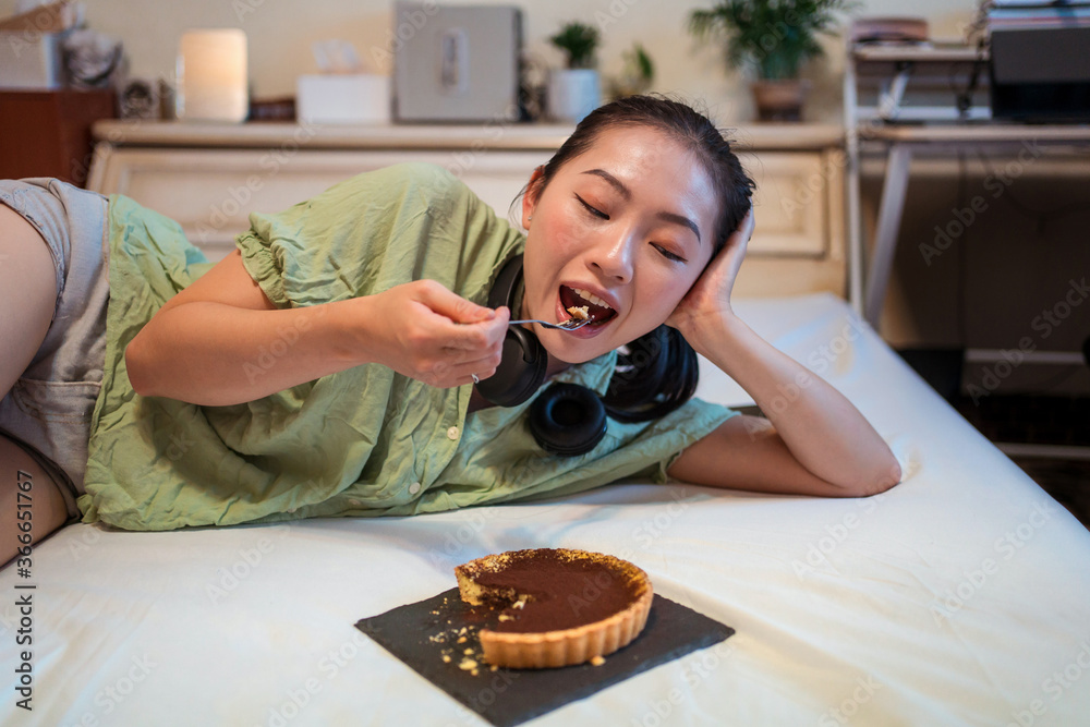 Young pretty Asian woman eating delicious chocolate tart lying on bed
