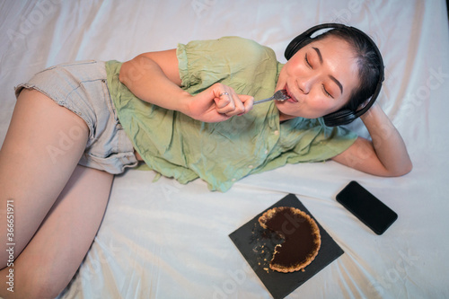 A top view of young pretty Asian woman eating delicious chocolate tart on bed
