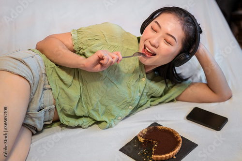 Obraz na płótnie Young pretty Asian woman eating delicious chocolate tart lying on bed licking fork while listening music w salonie