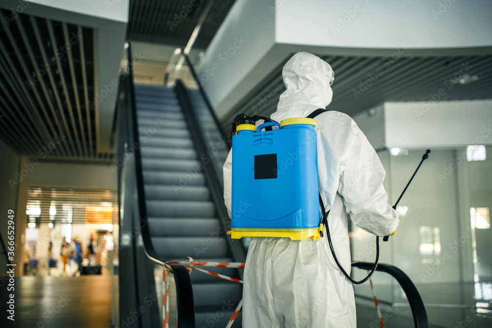 Portrait of a man in a sanitizing disifection suit holding spray near the escalator in an empty shopping mall. A volunteer cleaning up the public places to prevent covid-19. Health awareness concept. Portrait of a man in a sanitizing disifection suit holding spray near the escalator in an empty shopping mall. A volunteer cleaning up the public places to prevent covid-19. Health awareness concept.