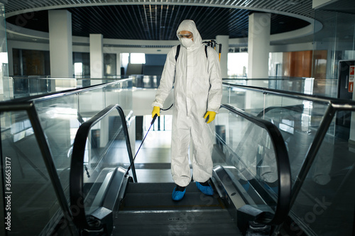 Disinfection worker wearing a protective suit professionally cleans up an escalator in an empty business center. A man equiped with antibacterial clothes sterilizes shopping mall. Covid concept.