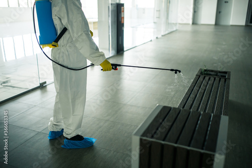 A man wearing protective suit is disinfecting a bench in an empty shopping mall with sanitizing spray. Cleaning up the public place to prevent covid spread. Healthcare precautions and safety concept. © Konstantin Zibert