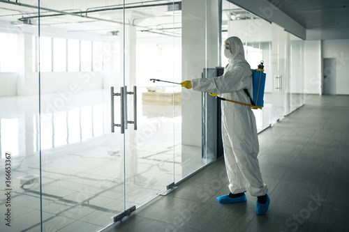 Sanitary worker sprays an empty business center with antiseptical liquid to prevent covid-19 spread. A man wearing disinfection suit cleaning up the shopping mall. Nobody, health, isolated concept.