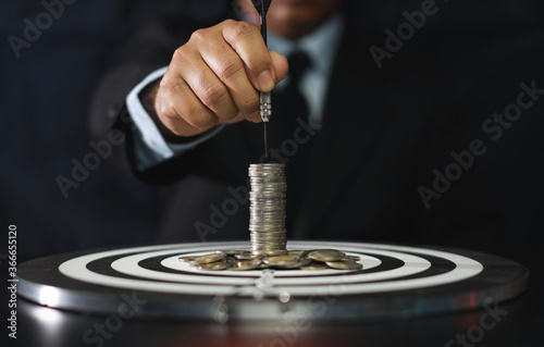 Businessman hands in black suite sitting and holding black dart put to centre of target board with stacking coin on vintage table meaning of investment and achievement goal. Plann and strategy concept