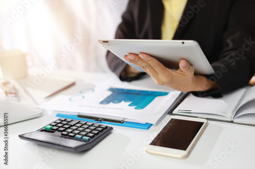 Businesswoman hands in black suit sitting and touching tablet black screen. Woman working or using computer on white table with business chart and calculator in modern office with paper work