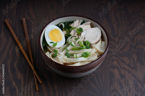 Japanese ramen soup made of chicken broth and noodles decorated with green onion, halved egg and meat slices in a bowl standing on dark brown wooden table together with chopsticks
