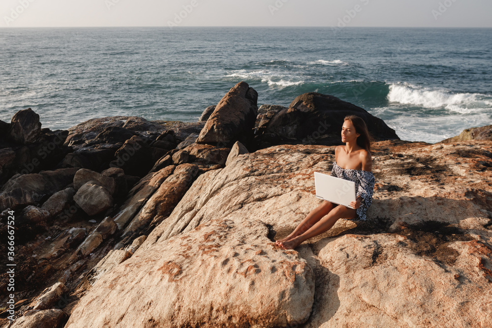 young woman using laptop on the beach, while sit on the rock with amazing ocean view. work and travel concept. Freelancer work online