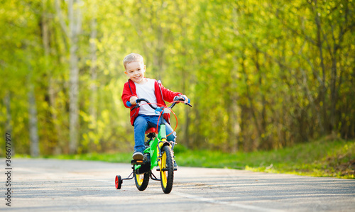 Boy kid training to ride bicycle with supporting wheels on sides, background summer park © Parilov