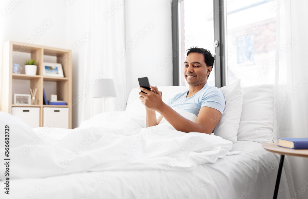 technology, bedtime and rest concept - happy smiling indian man with smartphone lying in bed at home technology, bedtime and rest concept - happy smiling indian man with smartphone lying in bed at home