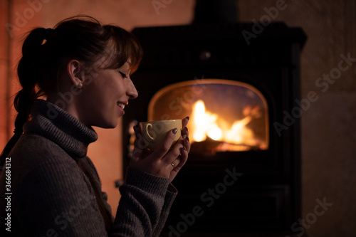 Portrait of young girl in a warm sweater drinking hot drink from mug by the fireplace. Cozy home Christmas evening by the fireplace in a wooden house. Relaxing by the fireplace after busy work day.