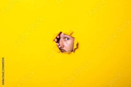 Young man looks with an eye into a torn hole in a yellow background