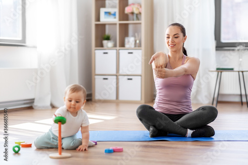family, sport and motherhood concept - happy smiling mother exercising on mat and little baby playing with toys at home