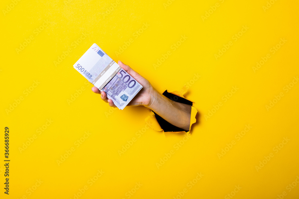 Woman's hand holds a bundle of five hundred euro bills on a yellow background. - obrazy, fototapety, plakaty 