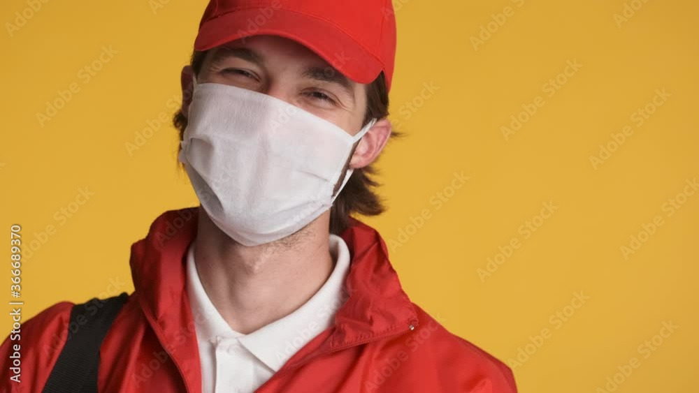 Close up attractive delivery guy in red cap and medical mask happily looking in camera over colorful background. Safety concept Close up attractive delivery guy in red cap and medical mask happily looking in camera over colorful background. Safety concept