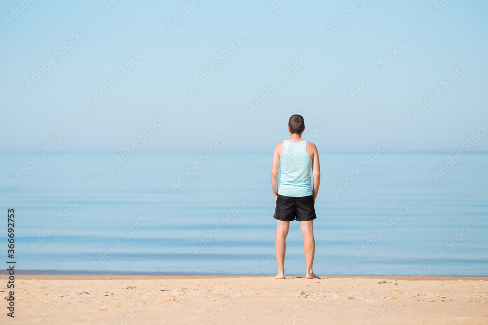 One young man standing alone on sand and staring at calm sea and light blue sky in warm summer morning. Empty place for emotional, positive, inspirational text, quote or sayings. Back view. One young man standing alone on sand and staring at calm sea and light blue sky in warm summer morning. Empty place for emotional, positive, inspirational text, quote or sayings. Back view.