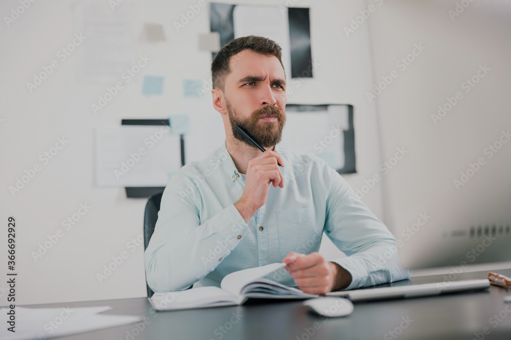young bearded brunette man looking thoughtful while working on business project in his modern office, holding pen in his hand, work routine concept young bearded brunette man looking thoughtful while working on business project in his modern office, holding pen in his hand, work routine concept